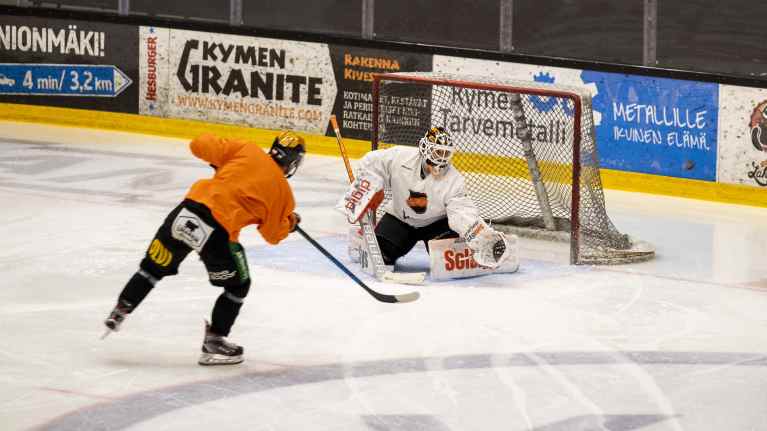 Oranssipaitainen jääkiekkoilija laukoo valkopaitaista maalivahtia kohti jääkiekkojoukkueen harjoituksissa.