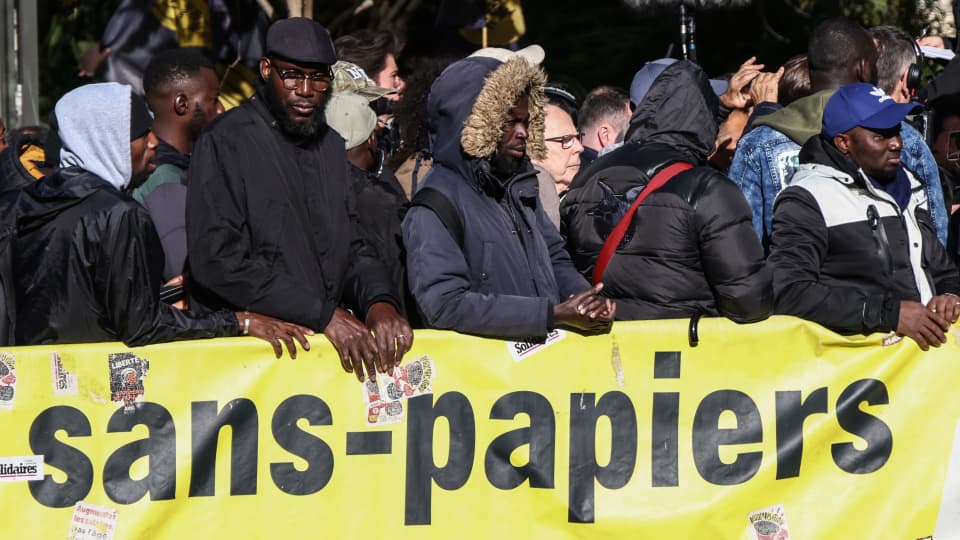 Mielenosoittajat seisovat ranskankielisen "paperittomat"-tekstin edessä.