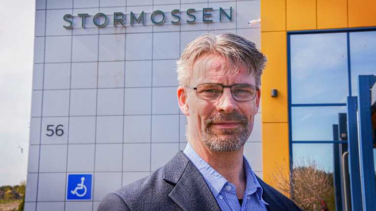 Aimo Latvala står utanför Stormossens byggnad. Han tittar in i kameran. Han är klädd i en grå kavaj och blå skjorta.