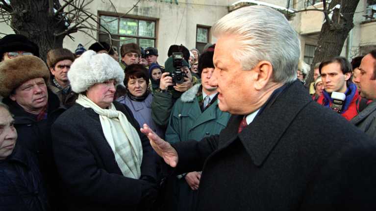 Boris Jeltsin seisoo etualalla tummassa päällystakissa ja puhuu kansalaisille kättään heiluttaen. Etualalla oleva nainen valkoisessa turkishatussa katsoo Jeltsiniä epäilevän näköisenä. Taustalla näkyy pankin seinää.