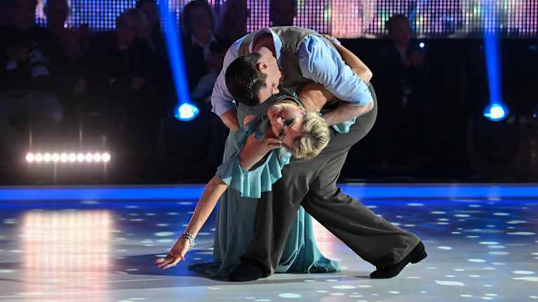 Sami Helenius taivuttaa Maria Guzeninaa tanssiessaan. Taustalla näkyy hämärässä istuva yleisö ja lavalle osottavat valonheittimet.