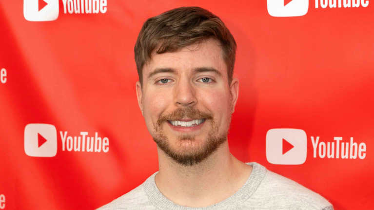 Ruskeahiuksinen nuori mies harmaassa villapaidassa hymyilee kameralle. Taustalla on Youtube-videopalvelun logoja.