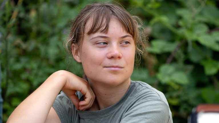 Greta Thunberg puolikuvassa, taustalla vihreitä pensaita ja puita.
