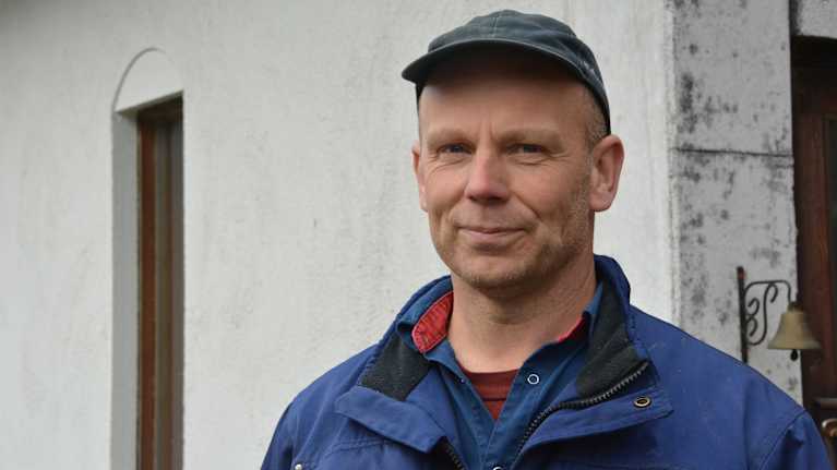 En man i keps och arbetskläder står framför sin ladugård, en vitrappad stenbyggnad.