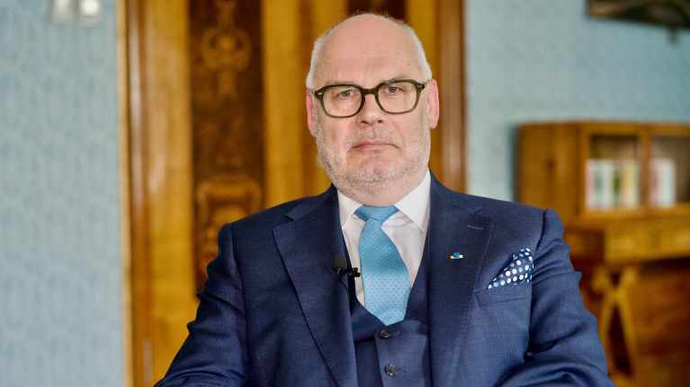 Silmälasipäinen pukuun ja solmioon pukeutunut mies katsoo suoraan kameraan.