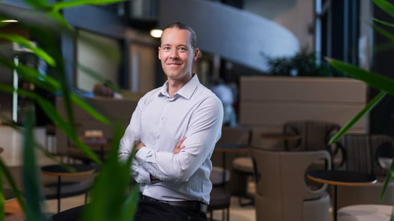 Mies hymyilee kameraan katsoen vaalea kauluspaita päällään kädet puuskassa.