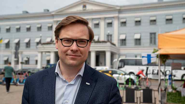 Tummansiniseen pikkutakkiin ja vaaleaan kauluspaitaan pukeutunut  Daniel Sazonov katsoo kameraan ja hymyilee. Taustalla näkyy kaupungintalo. 