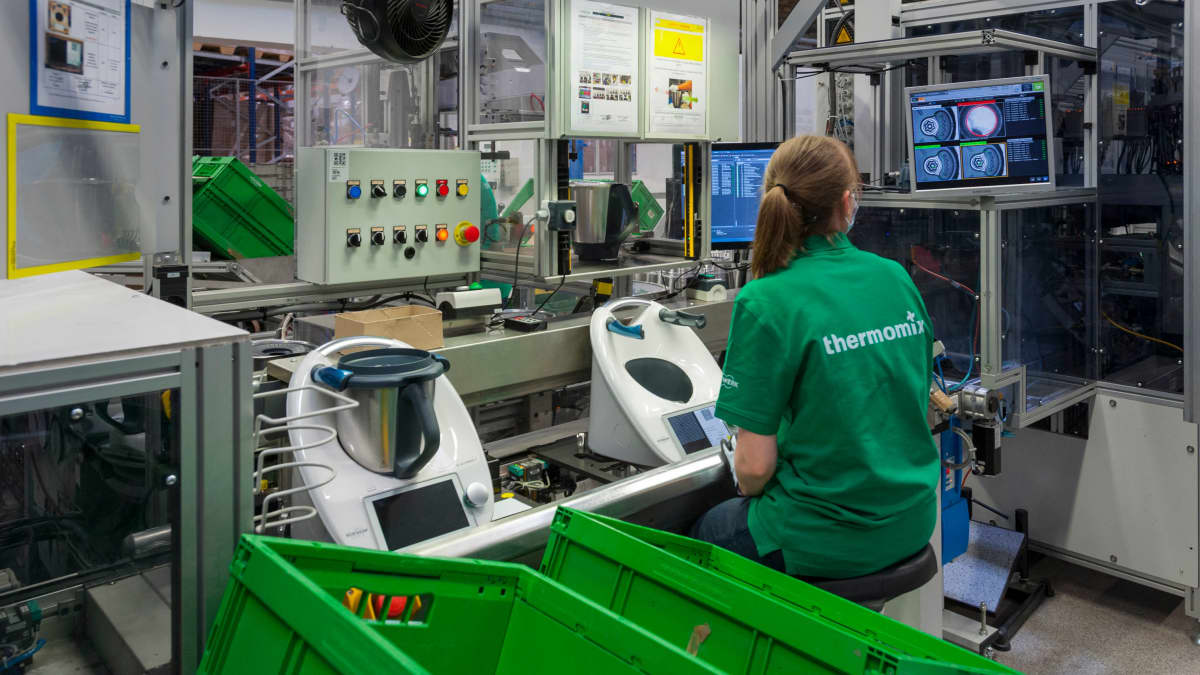 Moderni tuotantolinja, jossa näkyy tietokonepäätteitä ja vihreitä muovilaatikoita. Konetta käyttävä työntekijä on selin, selässä lukee Thermomix.