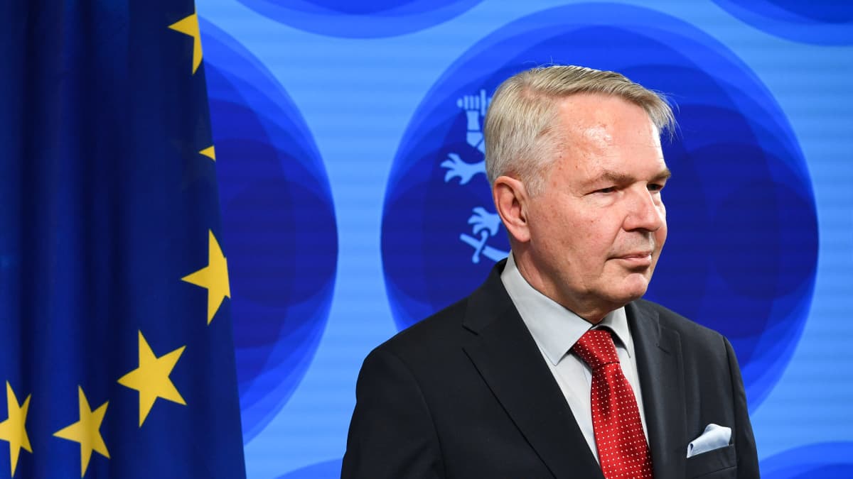 Man with grey hair, black blazer and red tie seen in semi-profile against wall with Finnish government logo and EU flag to the left.
