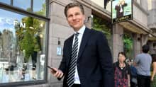 Antti Häkkänen i kostym håller i en telefon medan han går ut ur en byggnad. Han ler och tittar förbi kameran.