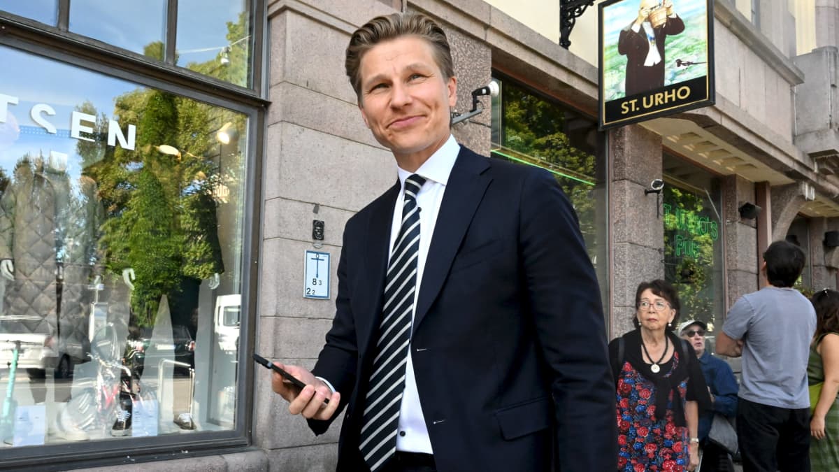 Antti Häkkänen i kostym håller i en telefon medan han går ut ur en byggnad. Han ler och tittar förbi kameran.