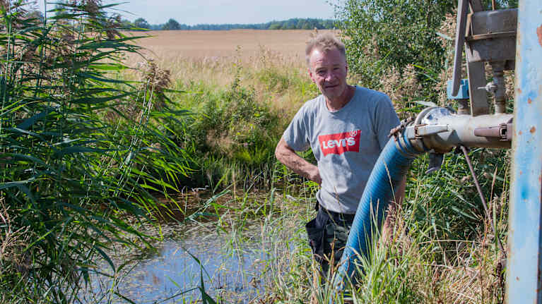 En jordbrukare står vid ett utfallsdike med vatten. Bredvid syns en slang som går till ett bevattningssystem.