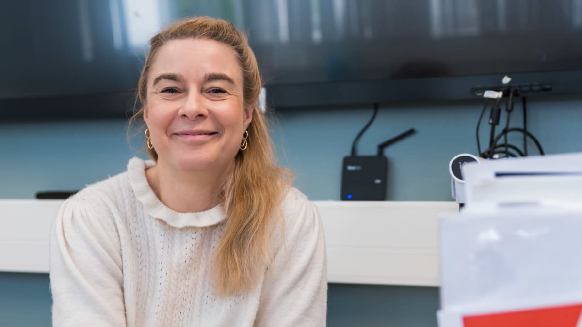 Nina Kurra istuu pöydän ääressä. Pöydällä tietokone ja kyniä purkissa sekä papereita.