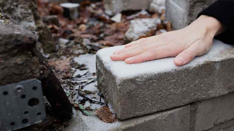 Käsi koskettaa ohutta lumikerrosta rauniotiilen päällä.