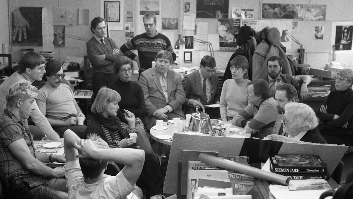 Mustavalkoinen kuva Helsingin Sanomien kulttuuritoimituksesta. Toimittajat istuvat kahvipöydän ääressä sotkuisessa toimituksessa 1980-luvun alkupuolella.
