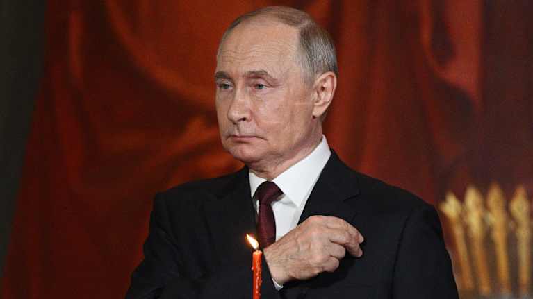 Vladimir Putin gör korstecknet. Ett tunt rött ljus brinner i förgrunden. 