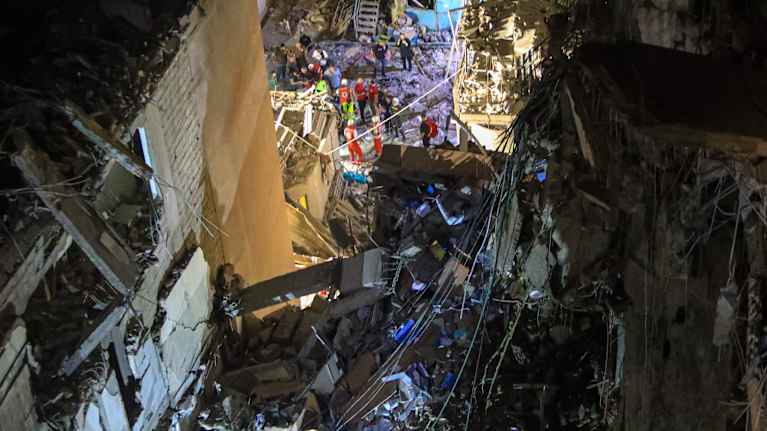 Rescue workers in orange overalls and others stand among the rubble of a building, seen from above.