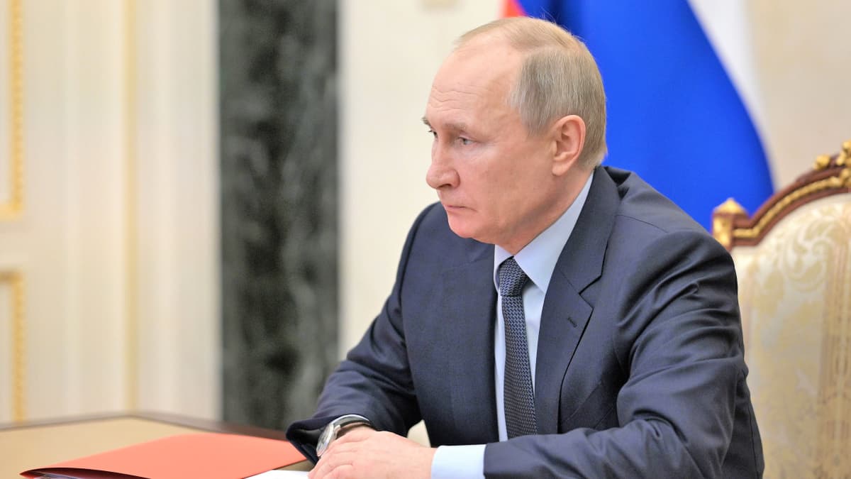 Putin istuu pöydän ääressä tummanharmaassa puvussa, vaaleansinisessä kauluspaidassa, tumma kravatti kaulassaan. Pöydällä edessä on pari paperia ja kynää ja oranssi kansio. Putinin ilme on vakava.