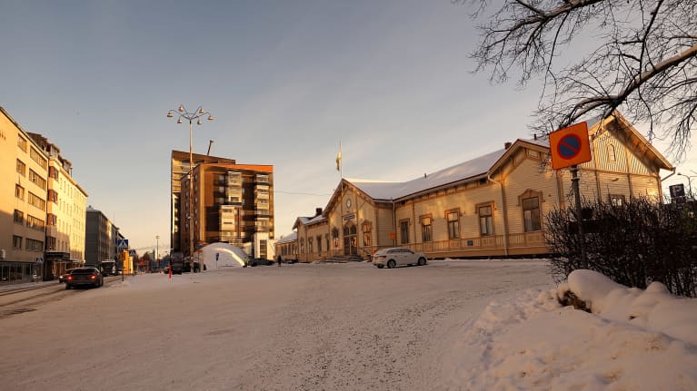 Talvinen kuapunkimaisema Oulun asema-aukiolta. Keltainen vanha VR:n puinen rautatieasema ja sen takana ruskea kerrostalo. Tien toisella puolella asemaa vastapäätä kerrotaloja ja autoja.