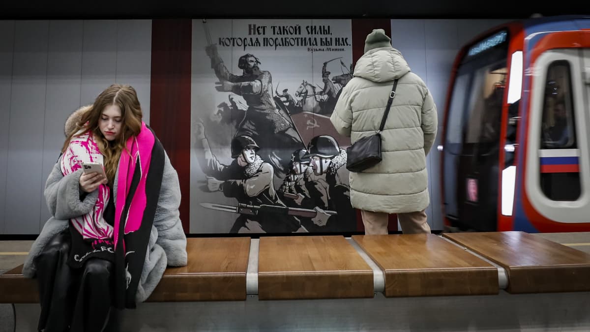 Metroasema, jossa Neuvostoarmeijaa ylistävä mainos seinällä. Mainoksen edessä seisoo ihminen ja metro on ajamassa laiturille. 