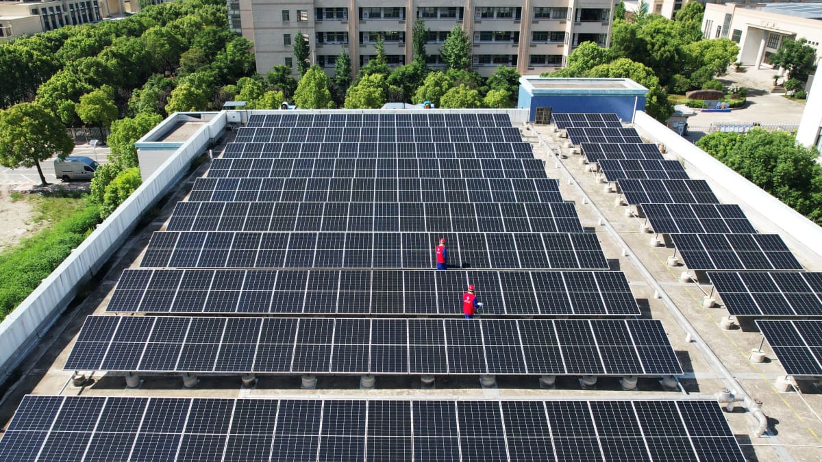 Kaksi henkilöä katolla, jolle on asennettu aurinkopaneeleita. Taustalla näkyy puistomainen kaupunkimaisema.