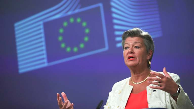 A woman with short grey hair speaks with her hands half raised in front of a blue background with the EU logo.