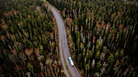 Aerial photo of a coach driving along a road surrounded by forest, with apartment buildings in the background