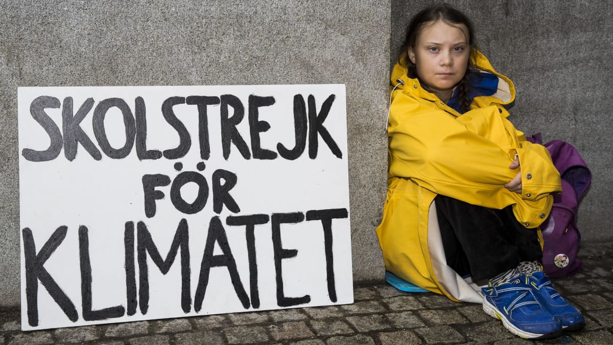 Greta Thunberg istuu seinää vasten. Hänellä on keltainen takki päällään ja vasemmalla puolella kyltti, jossa lukee ruotsiksi: "Koululakko ilmaston puolesta".