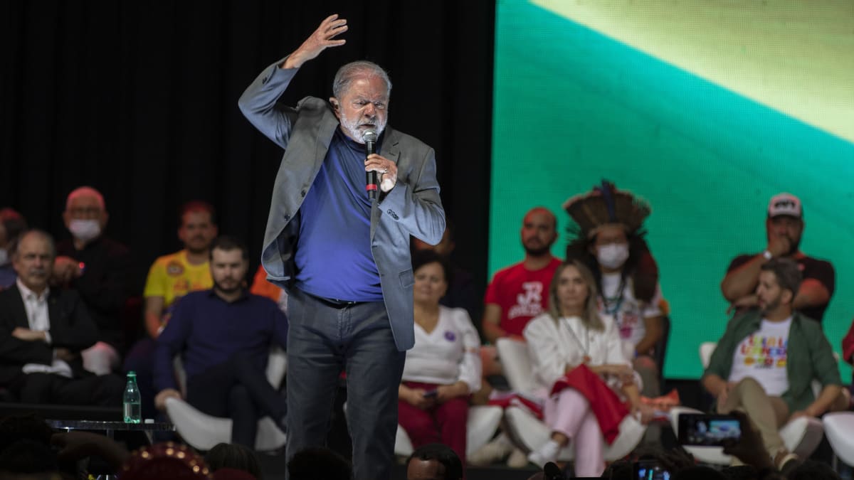Luiz Inácio Lula da Silva står på en scen och talar och gestikulerar. Han har en mikrofon i ena handen och den andra högt över huvudet. Han är klädd i grå kavaj och klarbå t-tröja.