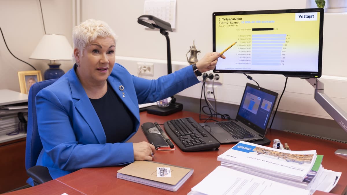 Siniseen bleiseriin pukeutunut nainen osoittaa tietokoneen näyttöä kynällä. Näytöllä on grafiikkaa. Naisen edessä olevalla ruskealla pöydällä on kannettava tietokone ja papereita.