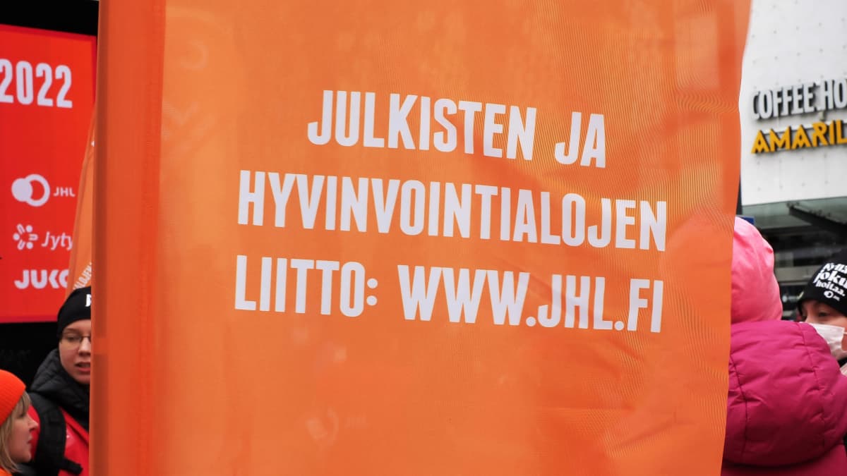 Oranssinen lippu, jossa valkoisella teksti JULKISTEN JA HYVINVOINTIALOJEN LIITTI: WWW.JUHL.FI. Taustalla ihmisiä.