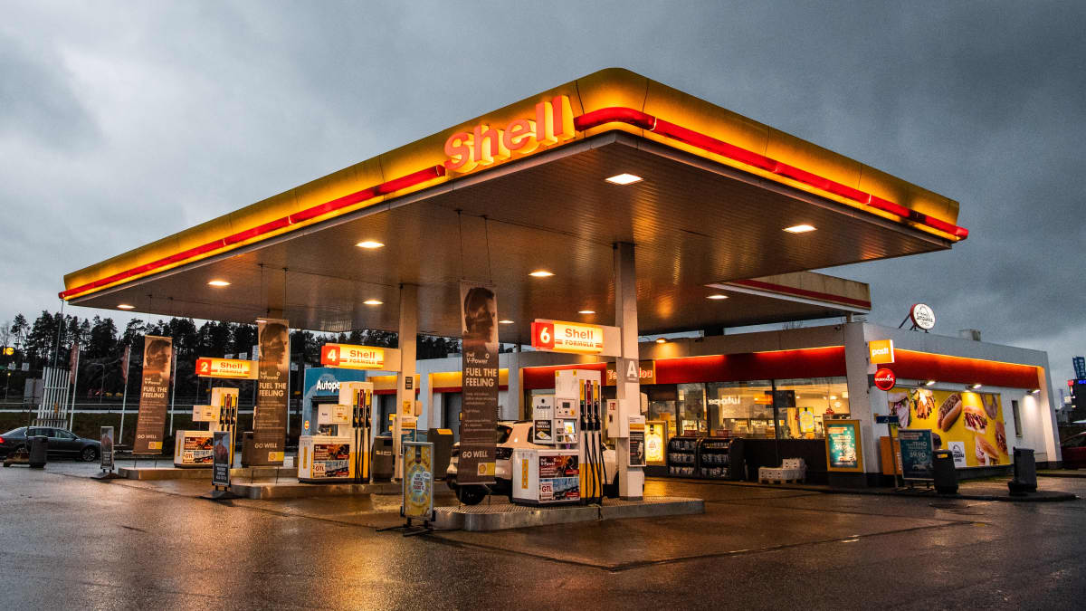 A Shell petrol station glows in yellow and red, beneath an overcast sky.