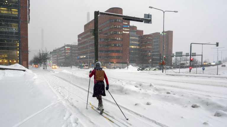 Nainen hiihtää reppu selässä Helsingissä jalkakäytävällä, sää on harmaa ja lunta runsaasti.