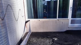Vaalean rakennuksen kulma, jota sotkettu spraymaalilla. Maahan kaivettu kuoppa ja seinästä revitty irti metallinen, pitkulainen osa.