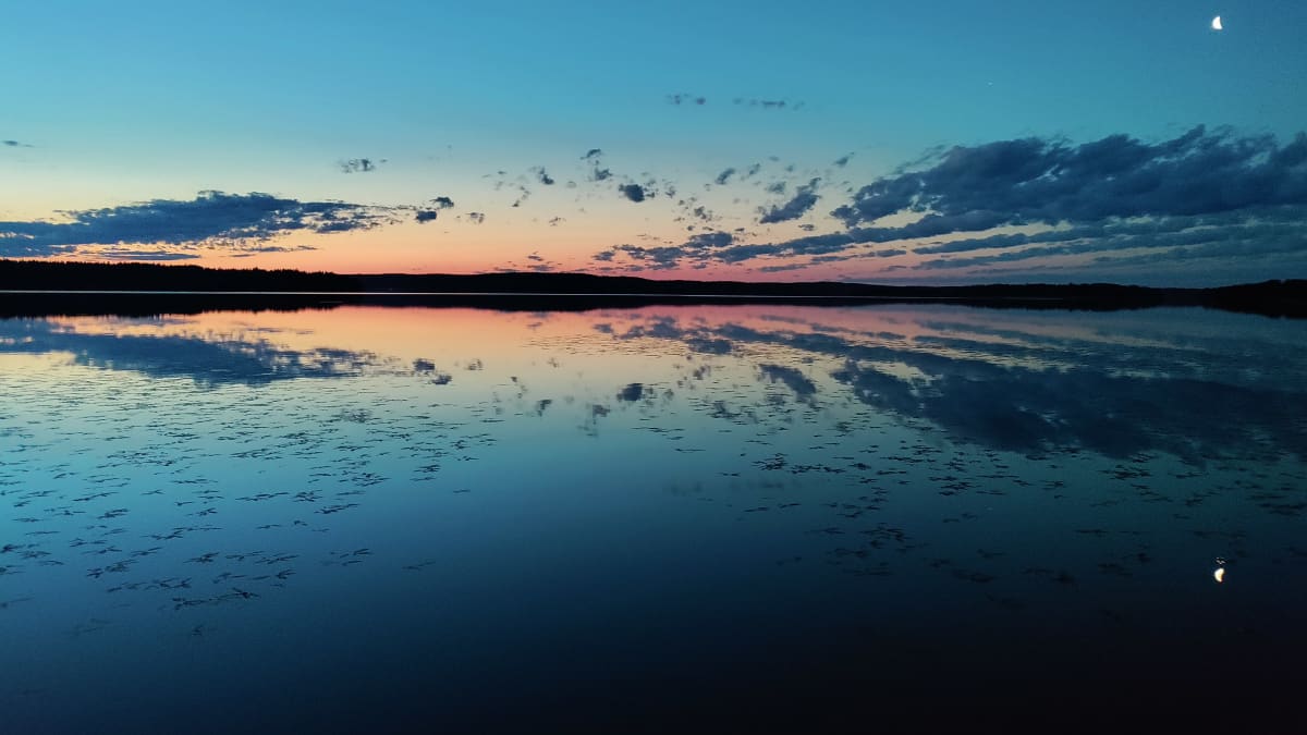 Aamun sarastus Vääksyssä. Aurinko nousemassa, tyyni vesi. Kuu vielä taivaalla. 