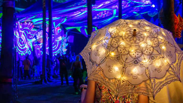 Henkilö seisoo valoilla koristellut aurinkovarjon alla ja katsoo piemässä hehkuvia taideinstallaatiota Komos Festivalilla.