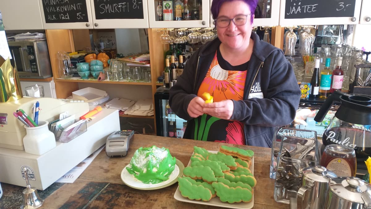 Hymyilevä nainen esittelee kirkkaan vihreää käpykakkua ja muita leivonnaisia.