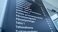 Musta sairaalan poliklinikkojen ja palvelupisteiden opastuskyltti, jossa valkoisella tekstillä eri osastojen nimiä ja lyhenteitä.