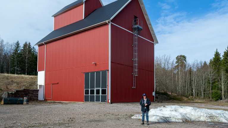 En stor röd lada med en person som står framför den på en grusbelagd mark. Omgivningen innehåller träd och en liten mängd snö på marken.