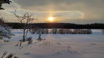 Aurinko pilkistää pilven aukosta. Lunta, peltoa ja metsää. 