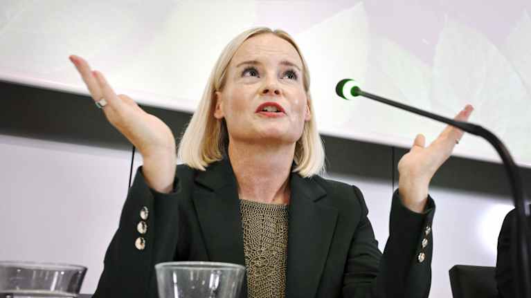 A blonde woman sits at a table, gesturing with her hands and speaking into a microphone, with a white wall in the background.