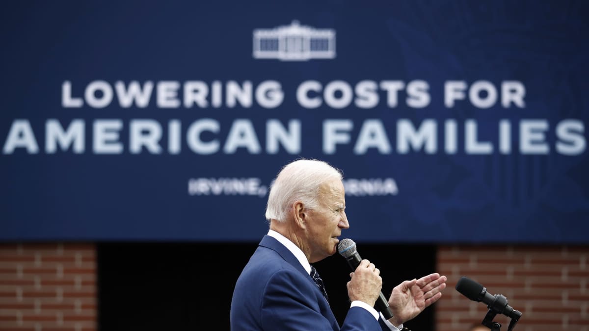 Harmaantunut vanha mies puhuu mikkiin ja osoittaa kädellään, sinisellä taustalla lukee "Lowering costs for American families".