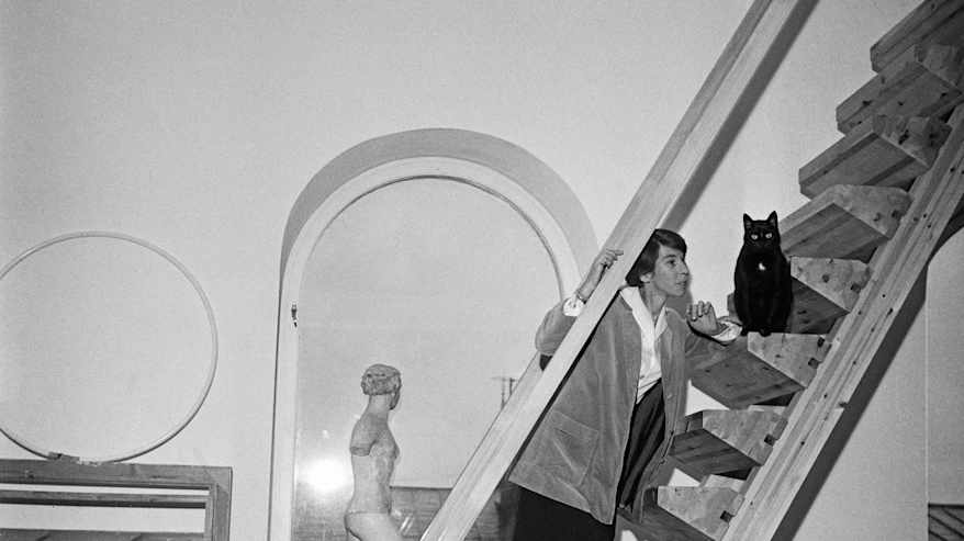 Mustavalkoisessa valokuvassa korkean ateljeetilan portaikossa poseeraa lyhythiuksinen henkilö väljässä takissa, hameessa ja korkokengissä, vieressään musta kissa.