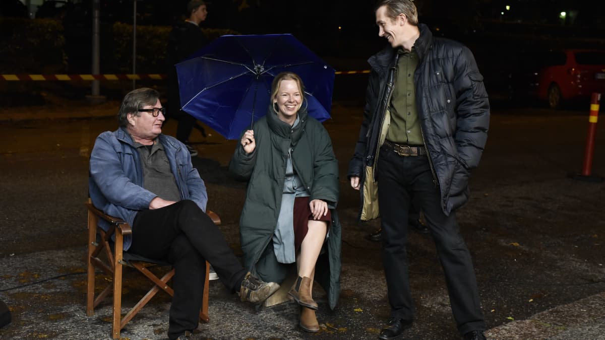 Director Aki Kaurismäki (seated at left) with actors Alma Pöysti (seated in the middle holding a blue umbrella) and Jussi Vatanen (standing on the right) on the set of Fallen Leaves.
