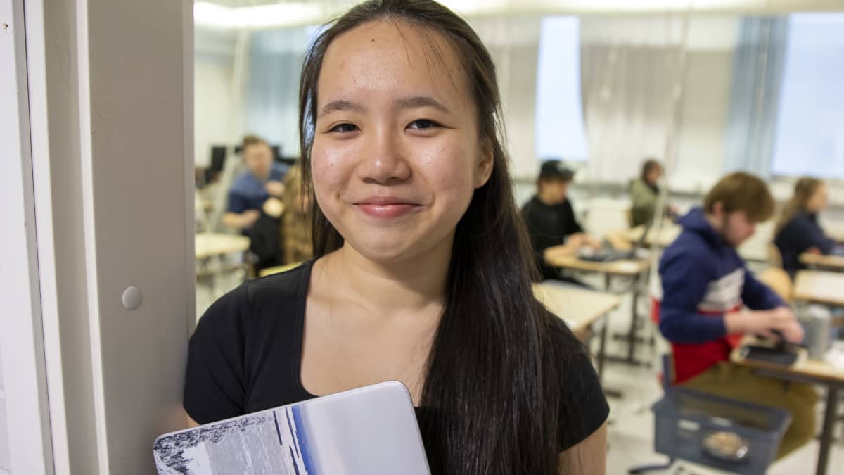 Thy "Anna" Nguyen seisoo luokkahuoneen ovella ja hymyilee kameralle. Taustalla näkyy yo-kokeen valmisteluhetkiä.