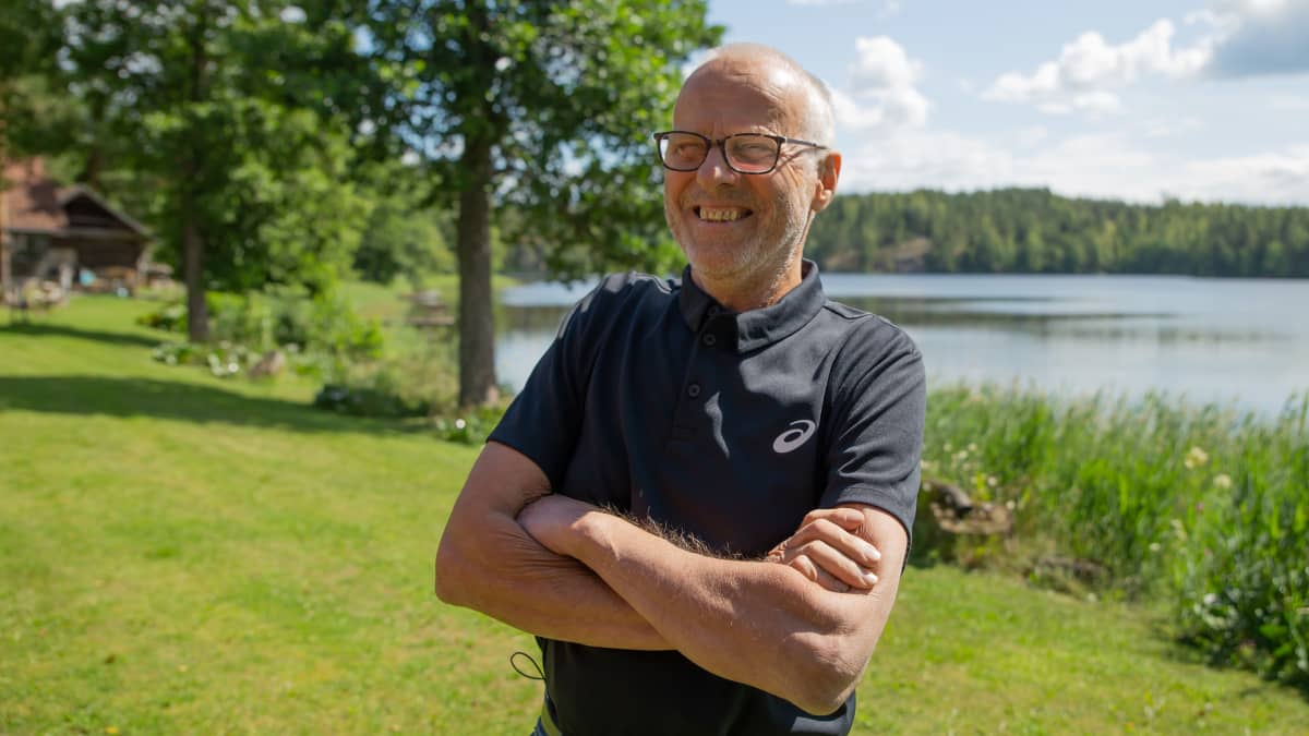 Lasse Viren iloisena kotitalonsa pihalla Myrskylässä. Aurinko paistaa, puut vihertävät ja järvi taustalla.