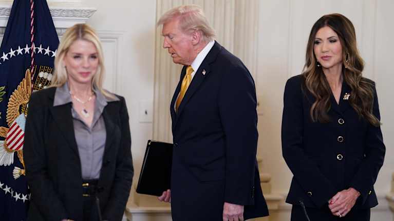 Pam Bondi ja Kristi Noem seisovat pöydän ääressä hymyillen jakkupuvuissa. Takana kulkee Donald Trump sinisessä puvussa ja keltaisessa kravatissa.