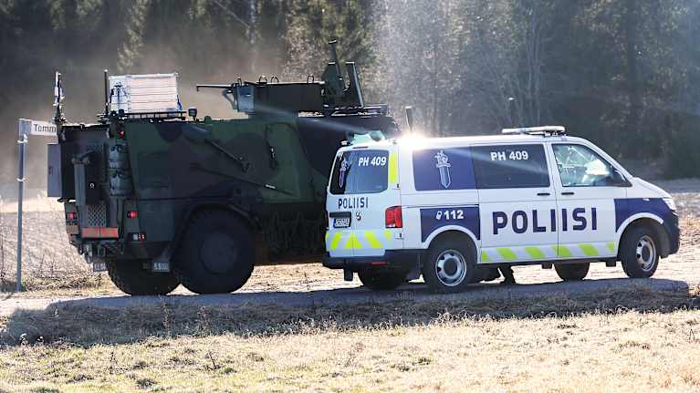 A green-and-black armoured vehicle and a blue-and-white police van parked on a sunny field with trees in the background.