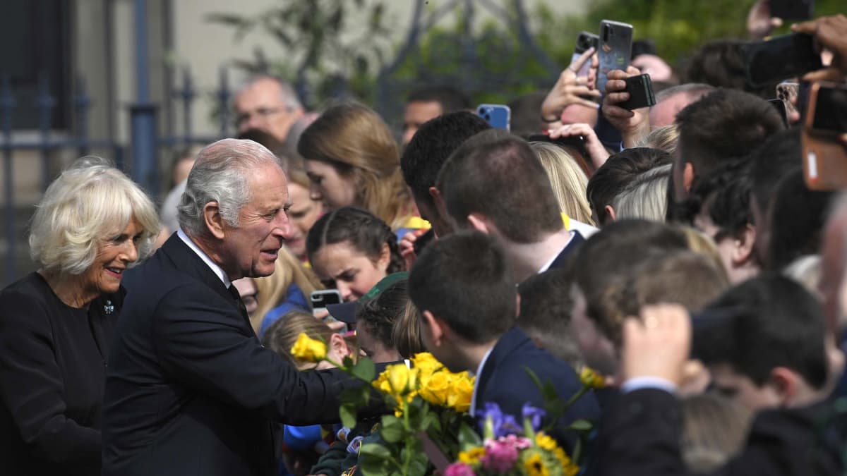Kung Charles och drottning Camilla, båda svartklädda, skakar hand med folk som står vid ett staket med blommor i händerna.