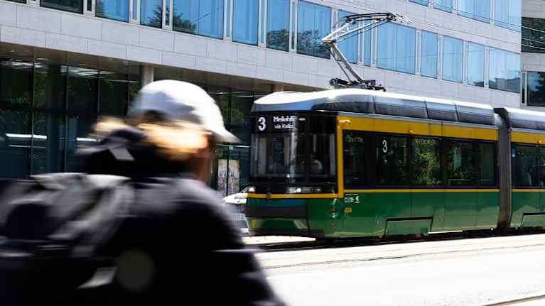 A blurry cyclist seen from behind and a green and yellow number 3 tram with an office building behind it.